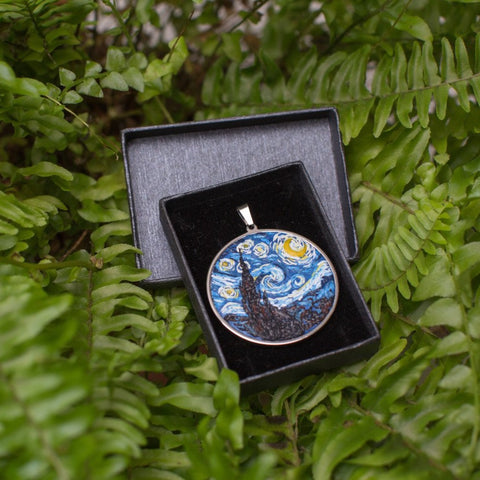 Round necklace which is made from stainless steel cabochon and filled with polymer clay to look like Van Gogh's painting Starry Nights. NEcklace is placed in black gift box and is in green fern background